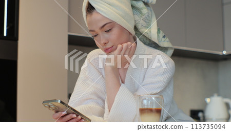 Portrait of happy woman wearing a bathrobe and a towel on her head using smartphone in the kitchen sending message on social media sharing. The morning is routine. Portrait of happy woman wearing a bathrobe and a towel on her head using smartphone in the kitchen sending message on social media sharing. The morning is routine. 113735094