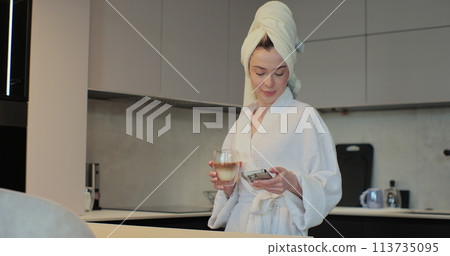 Beautiful young woman wearing a bathrobe and a towel on her head is drinking coffee and using smartphone in the kitchen. The morning is routine. Beautiful young woman wearing a bathrobe and a towel on her head is drinking coffee and using smartphone in the kitchen. The morning is routine. 113735095