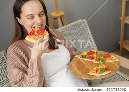 Joyful happy pregnant woman eating pizza alone at home female satisfied with timely food delivery sitting on sofa in living room enjoying fast food during pregnancy 113735098