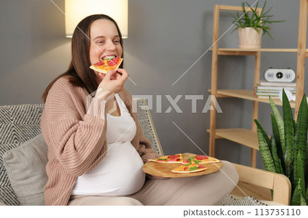 Cheerful satisfied pregnant woman biting big slice of pizza enjoying unhealthy nutrition relaxing at home with delicious fast food dinner eating with happy joyful face 113735110
