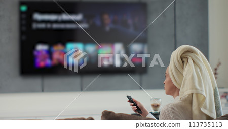 Young woman after a shower with towel on her head selects tv channels with remote control at living room on sofa. Woman controls TV using a modern remote control. Young woman after a shower with towel on her head selects tv channels with remote control at living room on sofa. Woman controls TV using a modern remote control. 113735113