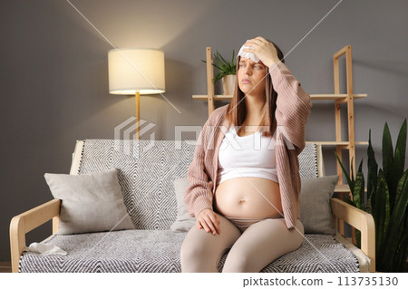 Young Caucasian pregnant woman with headache sitting on sofa couch in living room pregnancy flu symptoms expecting mother feeling hurt at home suffering high temperature touching painful forehead Young Caucasian pregnant woman with headache sitting on sofa couch in living room pregnancy flu symptoms expecting mother feeling hurt at home suffering high temperature touching painful forehead 113735130