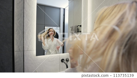 Young woman in bathrobe drying hair with a hairdryer in the bathroom at home. Usual morning beauty treatments in the bathroom. Young woman in bathrobe drying hair with a hairdryer in the bathroom at home. Usual morning beauty treatments in the bathroom. 113735139