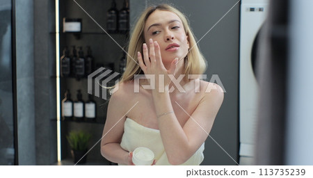 Happy young woman apply skincare cream on face look in bathroom mirror. Facial cream, healthy skin care treatment concept. 113735239