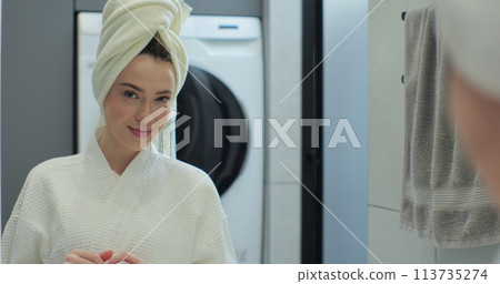 Portrait of young positive woman wearing bathrobe and towel on head after shower smiling to camera with mirror reflection. Natural make up, facial skincare. Portrait of young positive woman wearing bathrobe and towel on head after shower smiling to camera with mirror reflection. Natural make up, facial skincare. 113735274