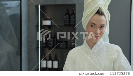 Portrait of young positive woman wearing bathrobe and towel on head after shower smiling to camera with mirror reflection. Natural make up, facial skincare. 113735276
