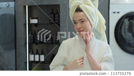 Portrait of young positive woman wearing bathrobe and towel on head after shower smiling to camera with mirror reflection. Natural make up, facial skincare. Portrait of young positive woman wearing bathrobe and towel on head after shower smiling to camera with mirror reflection. Natural make up, facial skincare. 113735278