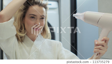 Close up of attractive young woman looking at her reflection in the mirror and drying hair with blow dryer in bathroom. Concept of well-groomed and beautiful woman. Close up of attractive young woman looking at her reflection in the mirror and drying hair with blow dryer in bathroom. Concept of well-groomed and beautiful woman. 113735279
