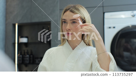 Attractive woman using a jade face roller in front of the bathroom mirror. Face massage. Beauty tool. Skin care. Attractive woman using a jade face roller in front of the bathroom mirror. Face massage. Beauty tool. Skin care. 113735297