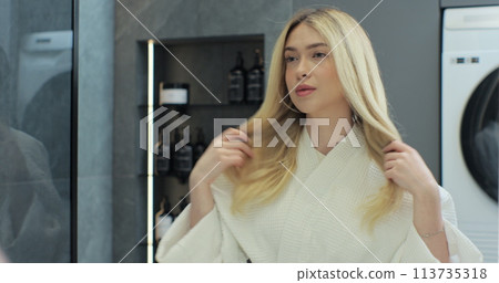 Woman looks into bathroom mirror touches her blond hair. Getting ready in morning, enjoying everyday routine. Wellness and organic skincare products. 113735318
