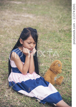 Sad girl hugging teddy bear sadness alone in green garden park. Lonely girl feeling sad unhappy sitting outdoors with best friend toy. Autism child play teddy bear best friend. Family violence concept 113741691