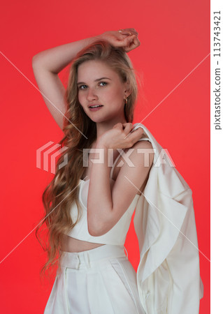 Beautiful woman of Caucasian ethnicity dressed in sculpting cupped corset top and jacket thrown over shoulder, is posing for camera on red background. She exudes positivity and represents Generation Z 113743421