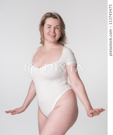Portrait of happy plump mature adult blonde woman in white body suit posing in studio on white background. Healthy lifestyle, body care concept. Smiling plus size Caucasian model looking at camera 113743473