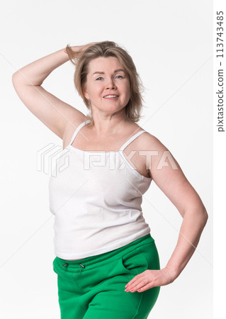 Portrait of happy plump mature adult blonde woman with medium length hair. Smiling model with one hand raised behind head wearing white cotton camisole and green sweatpants, posing on white background 113743485