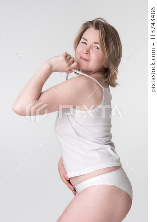 Woman wearing underwear holding strap of camisole and placing hand on stomach. Representative of concept of healthy lifestyle for women of Generation X Caucasian demographic, regardless of body type Woman wearing underwear holding strap of camisole and placing hand on stomach. Representative of concept of healthy lifestyle for women of Generation X Caucasian demographic, regardless of body type 113743486