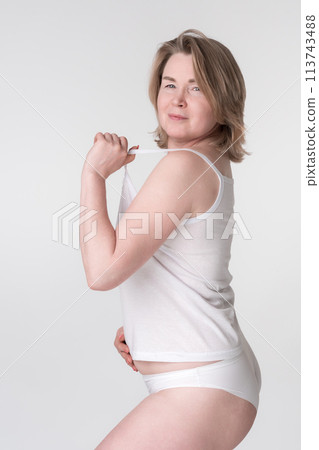 Middle aged overweight woman wearing white underwear, holding strap of camisole and placing hand on her stomach. Caucasian Generation X woman represents the concept of a healthy lifestyle for ladies 113743488