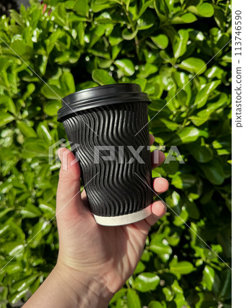 Hand holding black corrugated cardboard coffee cup with lid against background of lush green leafy bush. Urban eco friendly lifestyle. Blank space for sticker or label. 113746590