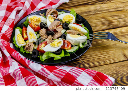 Tasty tuna salad with lettuce, black olives, eggs and fresh vegetables on wooden table Tasty tuna salad with lettuce, black olives, eggs and fresh vegetables on wooden table 113747409