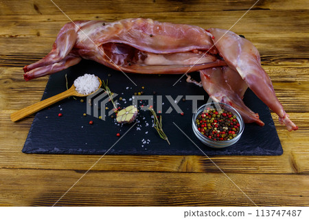 Whole raw rabbit meat with spices on a black slate cutting board on wooden table 113747487