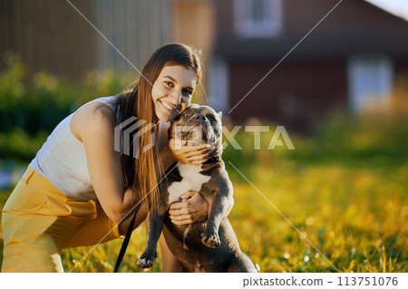 Trying to force dog to look into camera lens, young white woman takes photo with an American Bully in backyard of country house on summer evening. Trying to force dog to look into camera lens, young white woman takes photo with an American Bully in backyard of country house on summer evening. 113751076