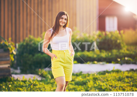 Knee length portrait of woman wearing yellow bermuda shorts, young white female stands against blurred rural background on summer evening before sunset. Knee length portrait of woman wearing yellow bermuda shorts, young white female stands against blurred rural background on summer evening before sunset. 113751078