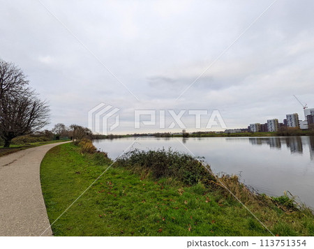 London, UK - 12.16.2021: An empty pathway next to Low Maynard Reservoir in the Walthamstow Wetlands along trees and marshes under a cloudy sky with buildings at the edge, viewed from Mayward Entrance 113751354
