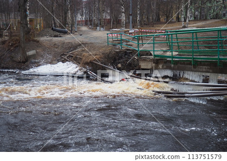 Spring flood. Rushing water in the river. Dark ferrous water rushes in the stream. Karelia, Lososinka River in spring. Floods, tsunami and climate change. Destroyed bridge with pipes. Weather disaster Spring flood. Rushing water in the river. Dark ferrous water rushes in the stream. Karelia, Lososinka River in spring. Floods, tsunami and climate change. Destroyed bridge with pipes. Weather disaster 113751579
