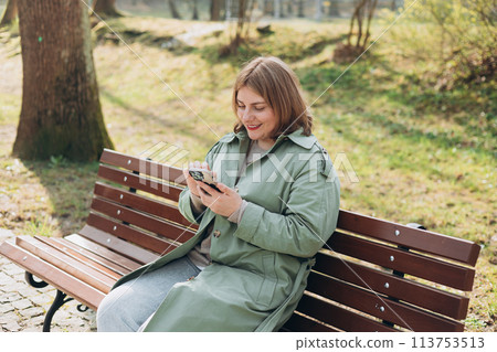 30s cheerful woman tourist dressed in casual look holding smartphone gadget in hand for communicate on the city street. Happy woman chatting on cell smartphone and relaxing on the bench outdoors 113753513