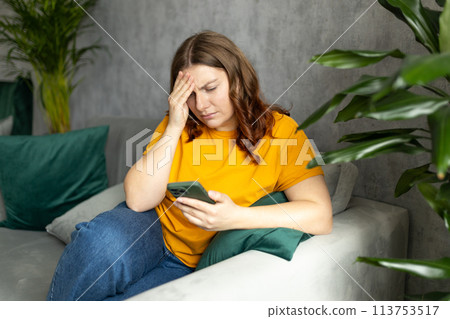 Portrait of beautiful young woman with depressed facial expression sitting on the couch holding her phone. Sad 30s women desperately looking at smartphone screen Portrait of beautiful young woman with depressed facial expression sitting on the couch holding her phone. Sad 30s women desperately looking at smartphone screen 113753517
