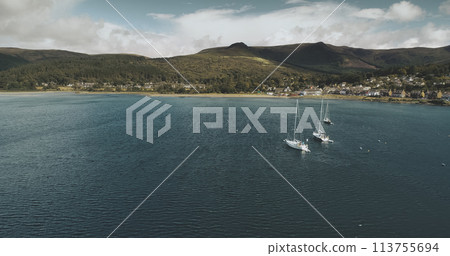 Atlantic ocean gulf, sailboats aerial zooming shot in Brodick Bay. Scottish landscape of port town. Houses, cottages, resort at shore of gulf against greenery lands and mountains view Atlantic ocean gulf, sailboats aerial zooming shot in Brodick Bay. Scottish landscape of port town. Houses, cottages, resort at shore of gulf against greenery lands and mountains view 113755694