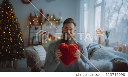 Person holding a red heart in a cozy room, decorated for Valentine's Day with a tree and lights. 113756098