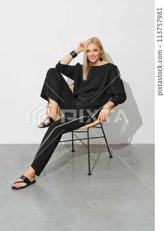 Smiling woman in loose casual clothes and leather sandals sits in profile on a chair Smiling woman in loose casual clothes and leather sandals sits in profile on a chair 113757981