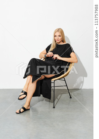 Cute woman in black sleeveless dress sits relaxed on a wicker chair 113757988