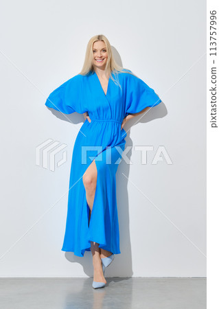 Smiling woman in vibrant blue dress leaning on white wall Smiling woman in vibrant blue dress leaning on white wall 113757996