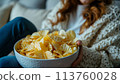 Bowl potato chips by the television. Modern TV set indoors, focus on bowl of chips. Female eating snack and watching tv. 113760028