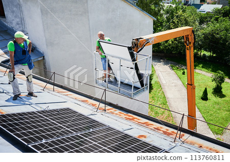 Workers lifting up photovoltaic solar panel on metal rooftop of house with assistance of crane lift. Men installers installing photovoltaic solar modules outdoors. Renewable energy generation concept. 113760811