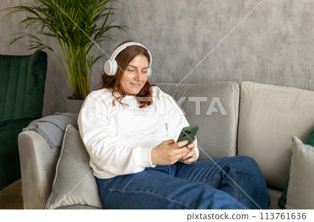 Smiling girl relaxing at home, she is listening to music, using a smartphone and wearing white headphones. Young 30s woman hearing enjoying good audio sound lounge on couch at home, full body Smiling girl relaxing at home, she is listening to music, using a smartphone and wearing white headphones. Young 30s woman hearing enjoying good audio sound lounge on couch at home, full body 113761636
