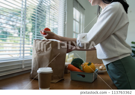 Organic Food Delivery. Happy young woman unpacking bag with Fresh Vegetables in kitchen Organic Food Delivery. Happy young woman unpacking bag with Fresh Vegetables in kitchen 113763362