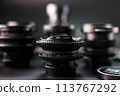 Selective focus on a vintage camera lens with serial number, surrounded by blur of other lenses, photography equipment array, black background setting 113767292