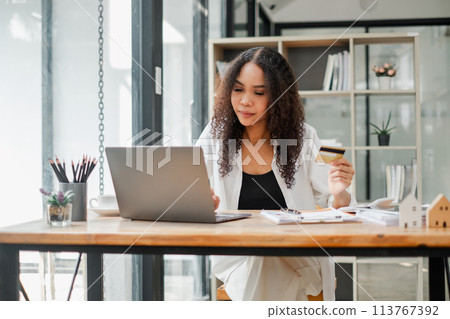 Businesswoman at a desk with a laptop, holding a credit card and looking intently at the screen. 113767392