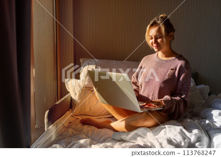 Happy casual beautiful woman working on a laptop sitting on the bed in the house. 113768247