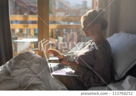 Happy casual beautiful woman working on a laptop sitting on the bed in the house. Happy casual beautiful woman working on a laptop sitting on the bed in the house. 113768252