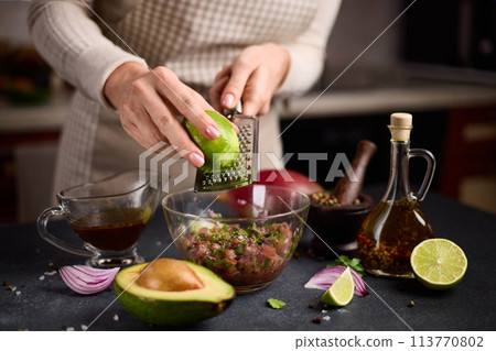 Woman grates lime zest onto chopped tuna, cilantro and onion in a glass bowl cooking traditional tuna tartare 113770802