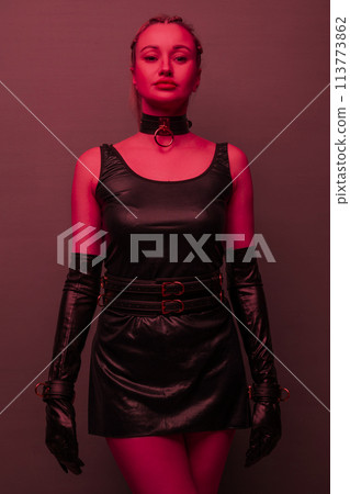Beautiful young woman in a leather dress and bondage set posing on red light backgound Beautiful young woman in a leather dress and bondage set posing on red light backgound 113773862