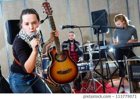 Attractive female soloist playing guitar and singing with her music band in sound studio 113779071