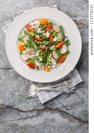 Spring salad of asparagus, radishes, cherry tomatoes, green peas and goat cheese close-up in a plate. Vertical top view 113779235