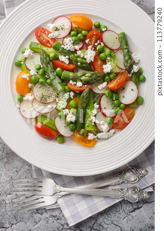 Light low-cadre salad of asparagus, fresh radishes, cherry tomatoes, green peas and goat cheese close-up in a plate on the table. Vertical top view 113779240