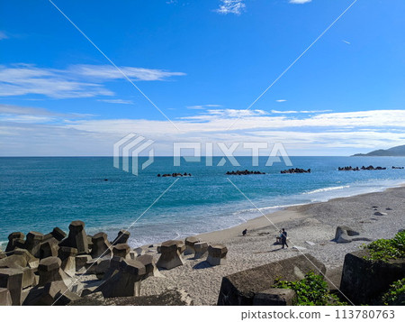 Hualien, Taiwan - 11.27.2022: People sitting and walking on a beach with tetrapod facing the Pacific Ocean under a sunny blue sky with mountains at the back during the pandemic 113780763