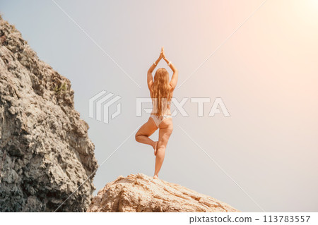 Woman sea yoga. Back view of free calm bliss satisfied woman with long hair standing on top rock with yoga position against of sky by the sea. Healthy lifestyle outdoors in nature, fitness concept. Woman sea yoga. Back view of free calm bliss satisfied woman with long hair standing on top rock with yoga position against of sky by the sea. Healthy lifestyle outdoors in nature, fitness concept. 113783557