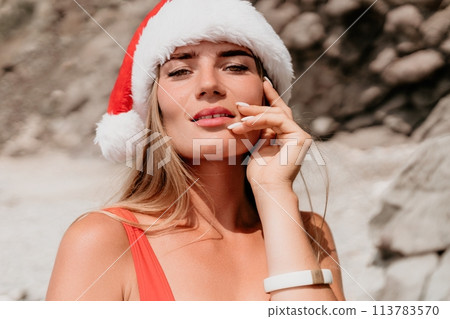 Woman summer travel sea. Happy tourist in red bikini and Santas hat enjoy taking picture outdoors for memories. Woman traveler posing on the beach surrounded by volcanic mountains, sharing travel joy 113783570